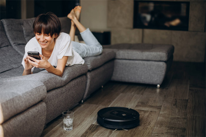 Девушка играет в телефоне лежа на диване пока робот-пылесос работает